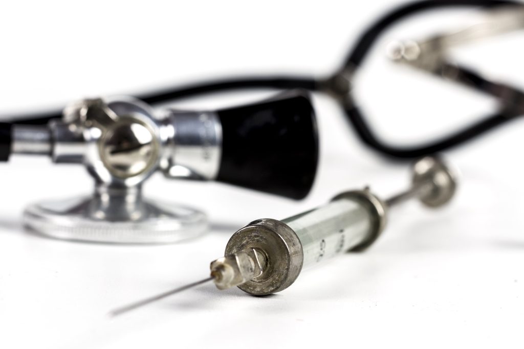 Syringe and stethoscope on a white surface, representing pharmaceutical production oversight